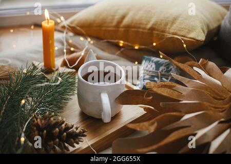 Warme Tasse Tee mit weihnachtslichtern, Sternen, Pinien, Kerzen und Kissen auf weichem Bett. Gemütliches Zuhause. Winterhygge. Festliche Weihnachtsdekorationen im Scan Stockfoto