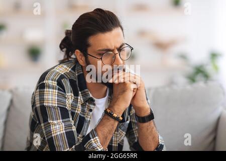 Nachdenklicher langhaariger indischer Kerl mit Brille, der zu Hause sitzt Stockfoto