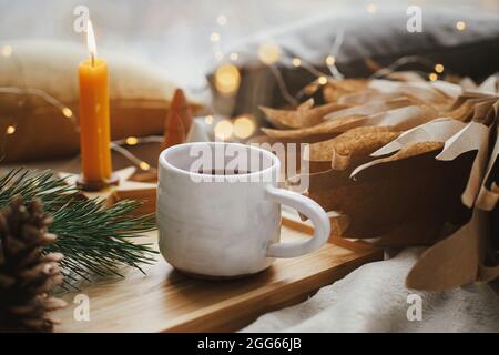 Winterhygge. Warme Tasse Tee mit weihnachtslichtern, Sternen, Pinien, Kerzen und Kissen auf weichem Bett. Gemütliches Zuhause. Festliche Weihnachtsdekorationen im Scan Stockfoto