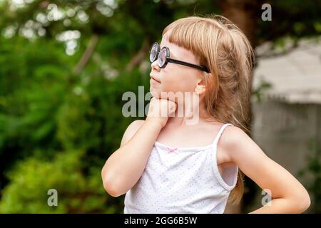 Ein kleines kluges Mädchen im jungen Schulalter, ein glückliches Kind mit einer großen, schrulligen Brille, das stolz steht und tief in Gedanken im Freien denkt Porträt, Stockfoto