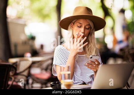 Junge Frau mit Hut beim Rauchen einer Zigarette und SMS. Modische Frau, die draußen auf der typisch französischen Café-Terrasse sitzt. Stockfoto
