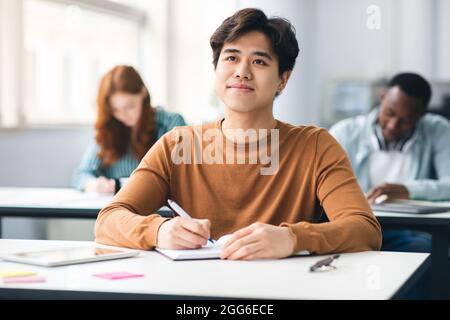 Lächelnder asiatischer Student, der im Klassenzimmer am Schreibtisch sitzt Stockfoto