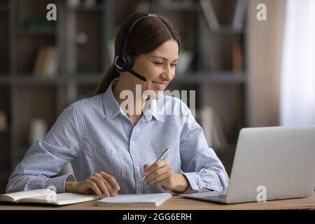 Lächelnde Frau mit Kopfhörern, die sich Notizen macht und zu Hause online studiert Stockfoto