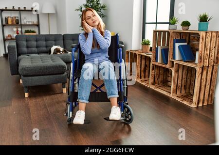 Junge schöne Frau, die zu Hause auf dem Rollstuhl sitzt und müde schläft und mit den Händen zusammen posiert, während sie mit geschlossenen Augen lächelt. Stockfoto