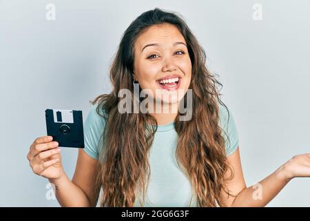 Junge hispanische Mädchen hält Diskette feiert Leistung mit glücklichen Lächeln und Sieger Ausdruck mit erhobener Hand Stockfoto