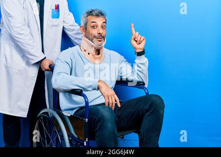 Ein hübscher Mann mittleren Alters mit grauen Haaren auf dem Rollstuhl, der einen Halsband trägt und mit dem Finger Nr. 1 nach oben zeigt, während er selbstbewusst lächelt Stockfoto