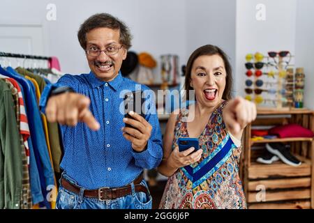 Interrassisches Paar im mittleren Alter im Einzelhandelsgeschäft mit einem Smartphone, das auf Sie zeigt, und der Kamera mit den Fingern, lächelnd positiv und fröhlich Stockfoto