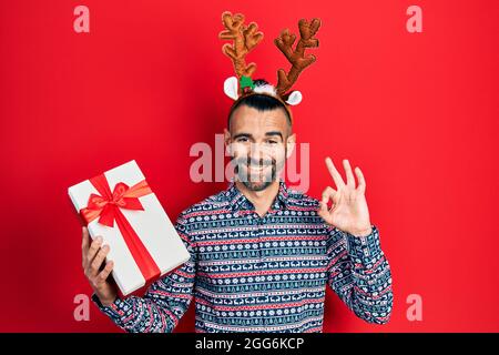 Junger hispanischer Mann trägt Hirsch weihnachtshut hält Geschenk tun ok Zeichen mit Fingern, lächelnd freundlich gestikulieren ausgezeichnetes Symbol Stockfoto