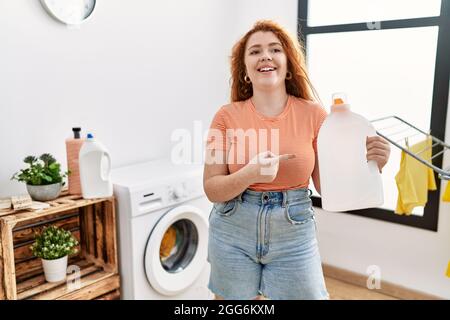Junge Rotschopf Frau macht Wäsche halten Waschmittelflasche lächelnd glücklich zeigt mit Hand und Finger Stockfoto