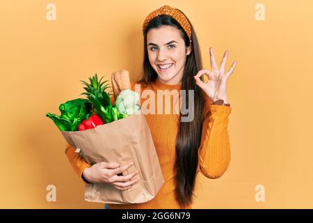 Junge Brünette Teenager halten Papiertüte mit Lebensmitteln lächelnd in der Liebe tun Herz Symbol Form mit Händen. Romantisches Konzept. Stockfoto