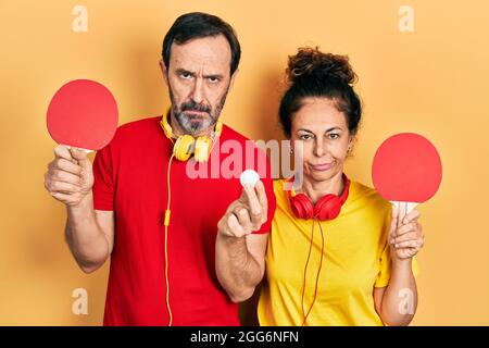 Mittelalterpaar hispanische Frau und Mann mit roten Ping-Pong-Schlägern skeptisch und nervös, runzelte die Stirn wegen des Problems. Negative Person. Stockfoto