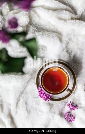 Hallo Frühling. Schwarzer Tee in einer Tasse mit einem kleinen Bouquet von Fliederblumen an der Seite auf einem weißen Wollbettdecke zu Hause. Draufsicht Stockfoto