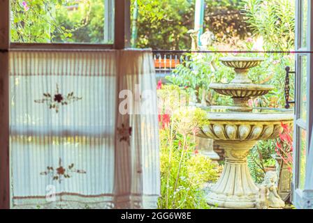 Handgefertigter weißer Vorhang mit Blick vom Fenster auf Brunnen und Grün aus Baumgarten Hintergrund Stockfoto