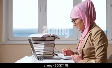 Eine muslimische Frau schreibt mit einem Stift in ein Notebook und schaut in einen Laptop Stockfoto