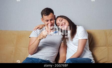 Eine junge Frau zeigte einem Mann, der auf der Couch saß, einen Schwangerschaftstest. Der Mann ist sehr glücklich, Vater zu werden Stockfoto