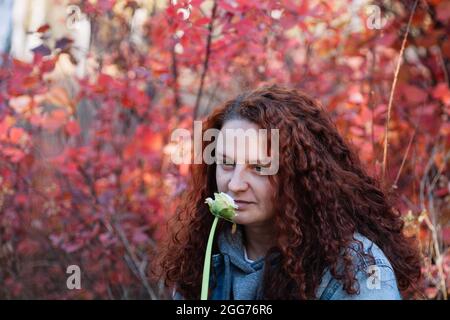 Frau mit Blume aus der Nähe. Frau mit langen kurvigen braunen Haaren riechen weiße Blume im Herbstwald mit rotem Busch auf dem Hintergrund Stockfoto