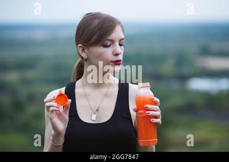 Frau trägt Sportkleidung und trinkt während des Trainings lebendigen Orangensaft. Abend- oder Morgenzeit in der Natur. Stockfoto