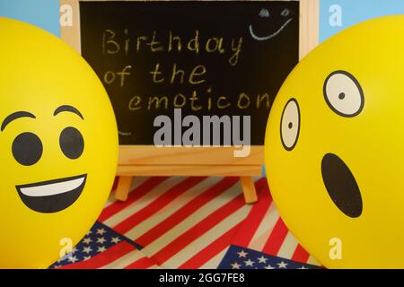 Gelbe Emoji-Ballons auf dem Hintergrund der Tafel mit der Ereignisbeschriftung und amerikanischen Flaggen. Stockfoto