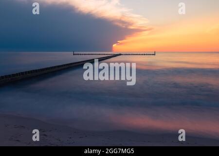 Untergehende Sonne vom Strand aus gesehen in Darłówko, Polen. Wunderschöne Meereslandschaft. Hölzerner Wellenbrecher am Horizont. Weichzeichnungseffekt (Langzeitbelichtung) Stockfoto