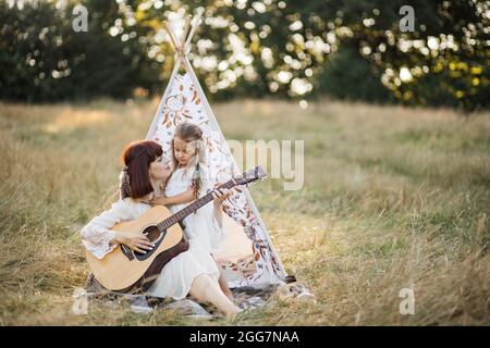 Gemeinsam Musik spielen und singen. Lächelnde, fürsorgliche Mutter lehrt ihre kleine Tochter, wie man Gitarre spielt und verbringt ihre Freizeit draußen auf dem Feld. Boho Frau und kleines Mädchen Spaß haben Stockfoto