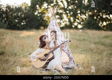 Happy stylish Boho Hippie Familie. Mutter und Tochter spielen Gitarre und singen zusammen. Erwachsene Attraktive Frau, die Gitarre für Mädchen spielt und im Sommerfeld vor dem Wigwam-Zelt sitzt Stockfoto