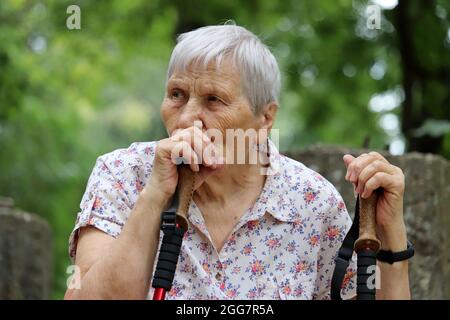 Nachdenkliche ältere Frau, die mit Spazierstöcken auf dem Hintergrund der Natur sitzt. Gesunde Lebensweise im Alter, Leben im Ruhestand Stockfoto