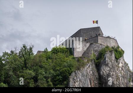 Dinant, Wallonien, Belgien - 8. August 2021: Zitadellenfestung gegen grauen Himmel mit belgischer Flagge und grünem Laub hoch über der Stadt. Stockfoto