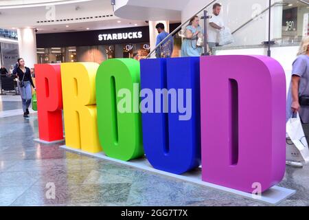 Die Menschen gehen an einem großen Schild mit der Aufschrift „PROUD“ vorbei, das in Regenbogenfarben für das Manchester LGBTQ Pride Weekend im Arndale Einkaufszentrum in Manchester, England, Stockfoto