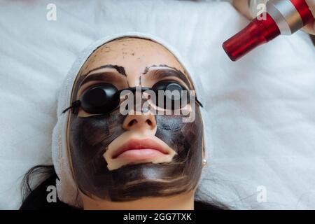 Nahaufnahme der Hand des Kosmetologen, die das Carbon Peeling der Hardware von einer jungen hübschen Kundin in einer Schutzbrille macht, die in Schönheit auf der Couch liegt Stockfoto