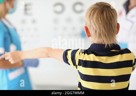 Der kleine Junge steht im Augenarztbüro vor den Ständen mit Buchstaben. Der Arzt prüft die Jungen auf Augenhöhe Stockfoto