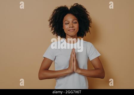 afroamerikanische junge Frau, die die Hände zusammengeklemmt hält und auf beigem Hintergrund betet Stockfoto