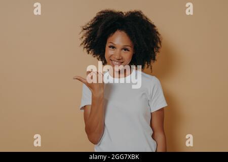 Junge lächelnde dunkelhäutige weibliche Lockenhaare zeigen mit Daumen zur Seite, isoliert über beige Wand Stockfoto