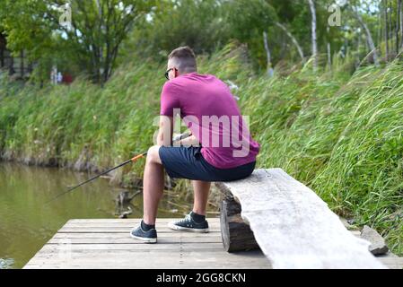 Junger Fischer sitzt auf hölzernen Pier, Angeln im See. Er hält die Rute und schaut auf Schwimmer Stockfoto