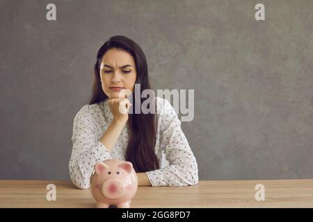 Frau, die mit einem Sparschwein an einem Tisch sitzt und plant, wofür sie ihr Geld ausgeben soll Stockfoto