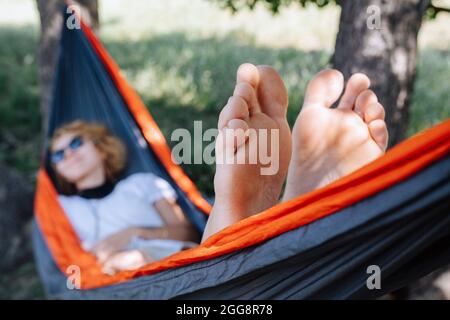 Eine Frau in Sonnenbrillen, die in einer Hängematte liegt und im Schatten zwischen zwei großen Bäumen schwungt. Konzentriere dich auf ihre Sohlen. Ruhe ist verschwommen Stockfoto