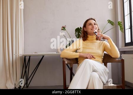Junge Frau in legerer Kleidung, die Mobiltelefon hält und glücklich träumt, während sie zu Hause auf dem Stuhl sitzt Stockfoto