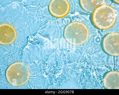 Zitronenscheiben in sauberem, transparentem Wasser auf blauem Hintergrund mit Kopierfläche. Wasser spritzt im Sonnenlicht auf die blaue Wasseroberfläche. Draufsicht oder Flatlay. Sommer, Urlaub, gesunde Ernährung Konzept Stockfoto