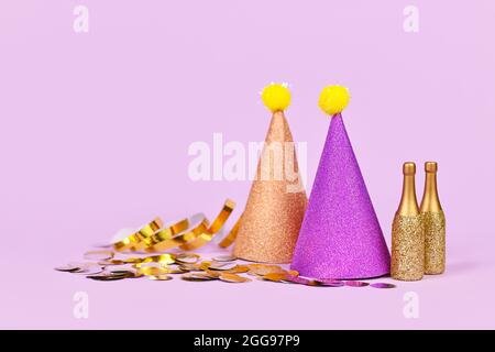 Glitzernder Partyhut, kleine goldene Champagnerflaschen und Partystreamer auf violettem Hintergrund mit Kopierraum Stockfoto
