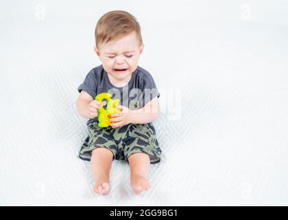 Trauriger Junge in einem khaki-Anzug mit buntem Rassel-Spielzeug, das auf dem Bett sitzt und weint. Porträt eines unglücklichen kaukasischen kleinen Kindes. Stockfoto