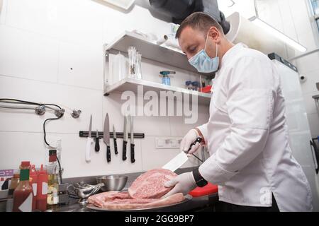 Der Restaurantchef trägt eine medizinische Gesichtsmaske und schneidet in der Küche Rindfleisch Stockfoto