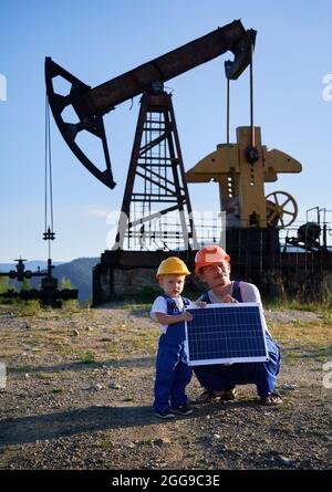 Spezialist in der Ölindustrie mit kleinen männlichen Kind halten Panel der Solarbatterie in den Händen gegen Stangenpumpe in Ölkompressorstation. Alternative Ökoenergiekonzeption. Stockfoto