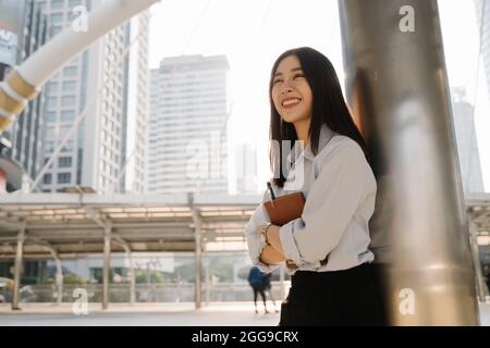 Fröhliche, schöne junge asiatische Frau, die sich an der Säule des Gebäudes lehnt und ein Notizbuch und einen Stift hält, während sie wegschaut Stockfoto