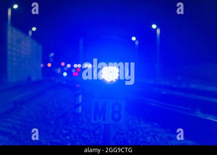 Die blaue Ampel am Nachtbahnhof leuchtet blau. In der Ferne Dunst und leuchtende Laternen. In der Nähe der Eisenbahn.verschwommener Hintergrund. Stockfoto