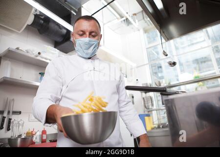 Aufnahme eines Küchenchefs mit medizinischer Maske, der im Restaurant Pommes frites zubereitet Stockfoto