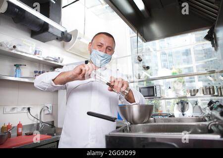 Fröhlicher Koch mit medizinischer Gesichtsmaske, salzender Teller in einer Schüssel in seiner Küche, Platz zum Kopieren Stockfoto