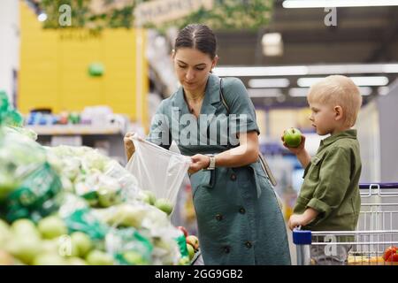 Junge Brünette Frau in Kleid setzen Äpfel in Plastiktüte, während ihr Sohn essen Apfel in den Warenkorb auf Bauernmarkt Stockfoto