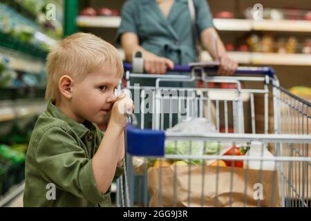 Müder kleiner blonder Junge in grünem Hemd, der sich auf den Einkaufswagen lehnt, während seine Mutter im Supermarkt Produkte kauft Stockfoto