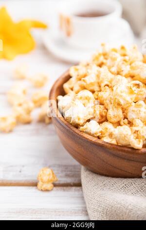 Popcorn mit Karamell in Holzschüssel und eine Tasse Kaffee auf weißem Holzhintergrund und Leinentextilien. Seitenansicht, Nahaufnahme, selektiver Fokus. Stockfoto