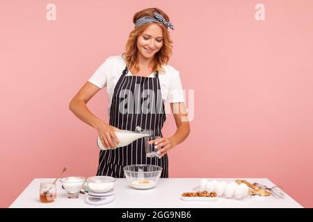 Junge Erwachsene Küchenchefin gießt die Milch ins Glas, Konditor in schwarzer Schürze knetet den Teig, bereitet Kuchen oder Kekse, hausgemachtes Gebäck zu. Indoor st Stockfoto