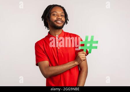 Abonnieren Sie den beliebten Blog! Bärtiger Mann mit Dreadlocks trägt ein rotes T-Shirt im lässigen Stil, hält ein Hashtag-Symbol für soziale Medien und lächelt. innenstudi Stockfoto